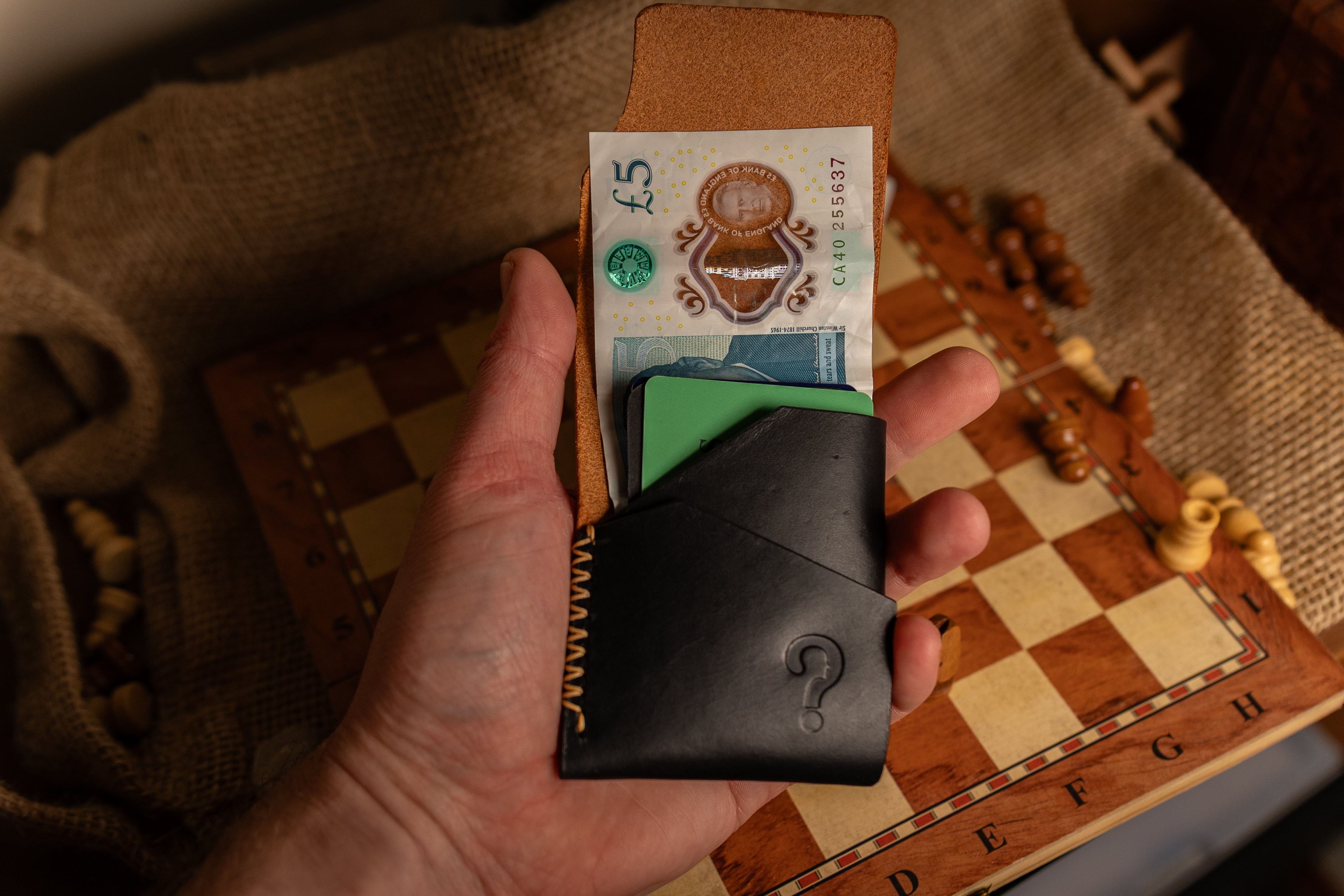 Hand holding a black leather wallet with a question mark design, displaying a few banknotes on a checkered fabric background.