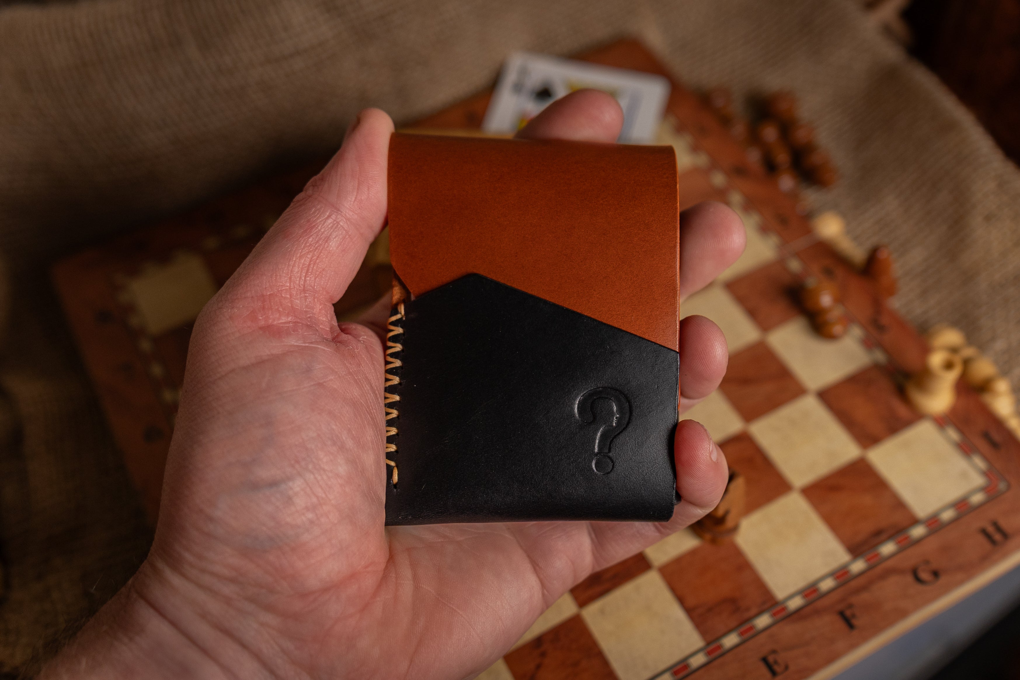 Hand holding a brown and black leather wallet with a question mark design on a checkered surface.