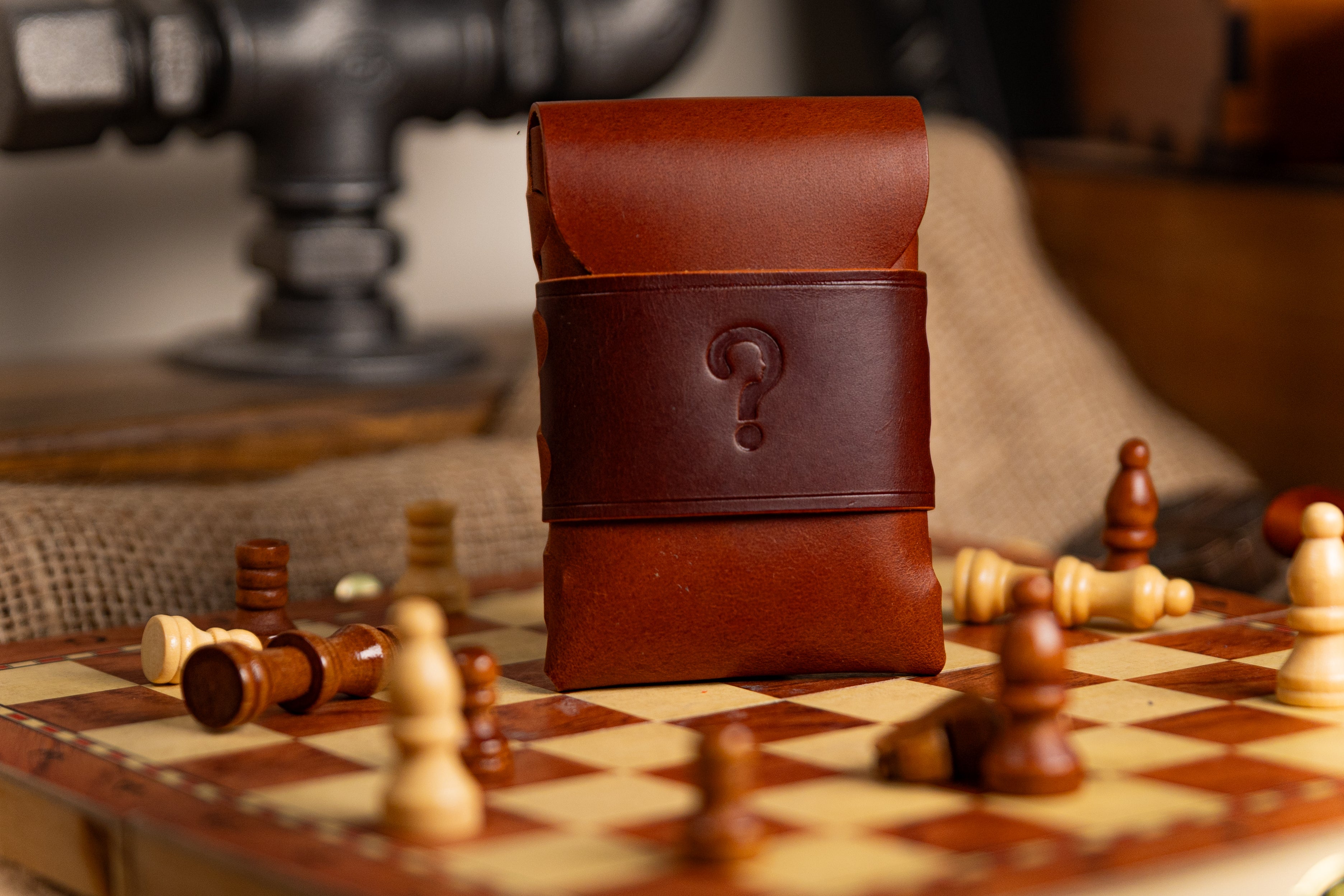 Brown walpier buttero leather pouch with a question mark on a chessboard with scattered chess pieces.