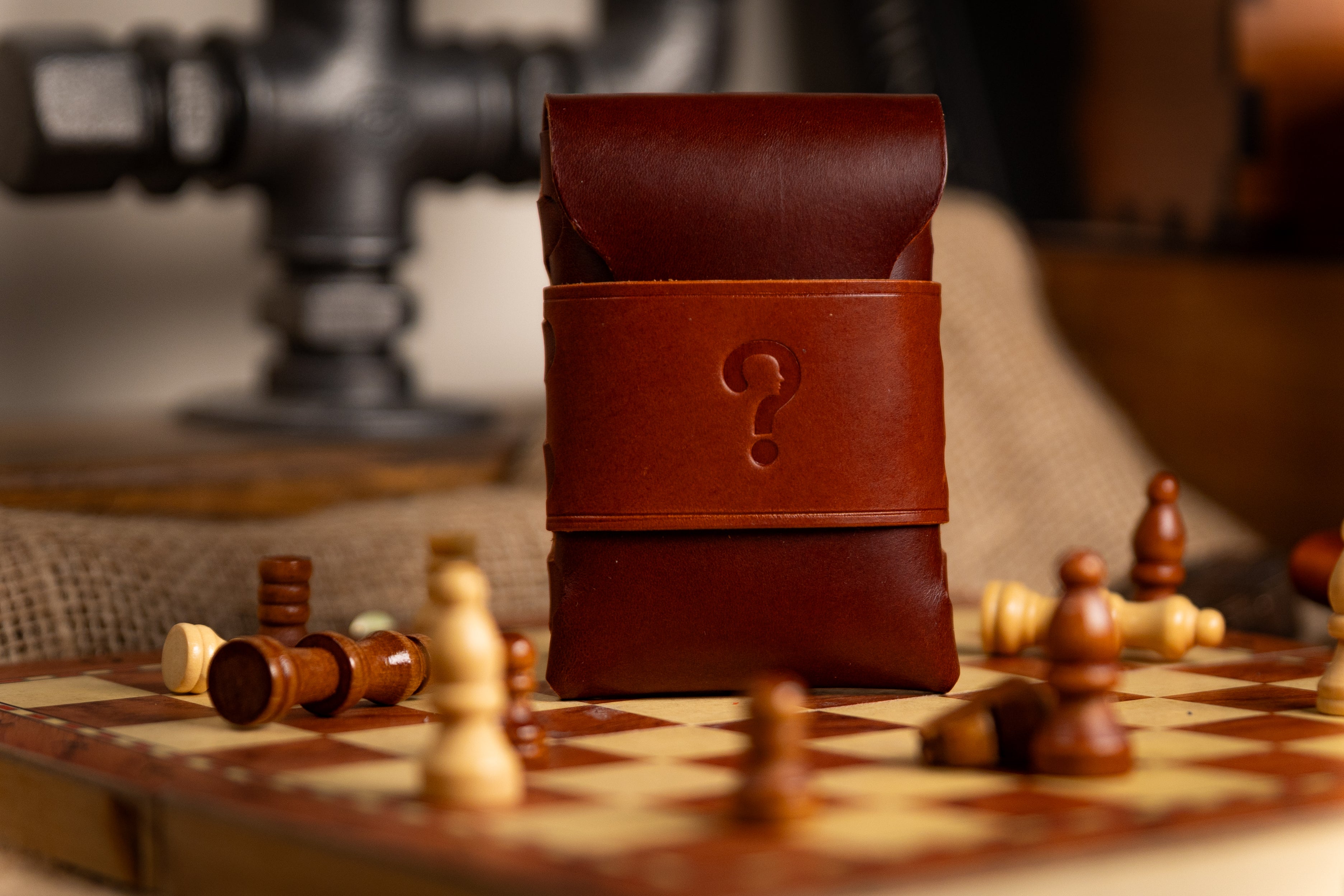 Brown walpier buttero leather pouch with a question mark on a chessboard with scattered chess pieces.