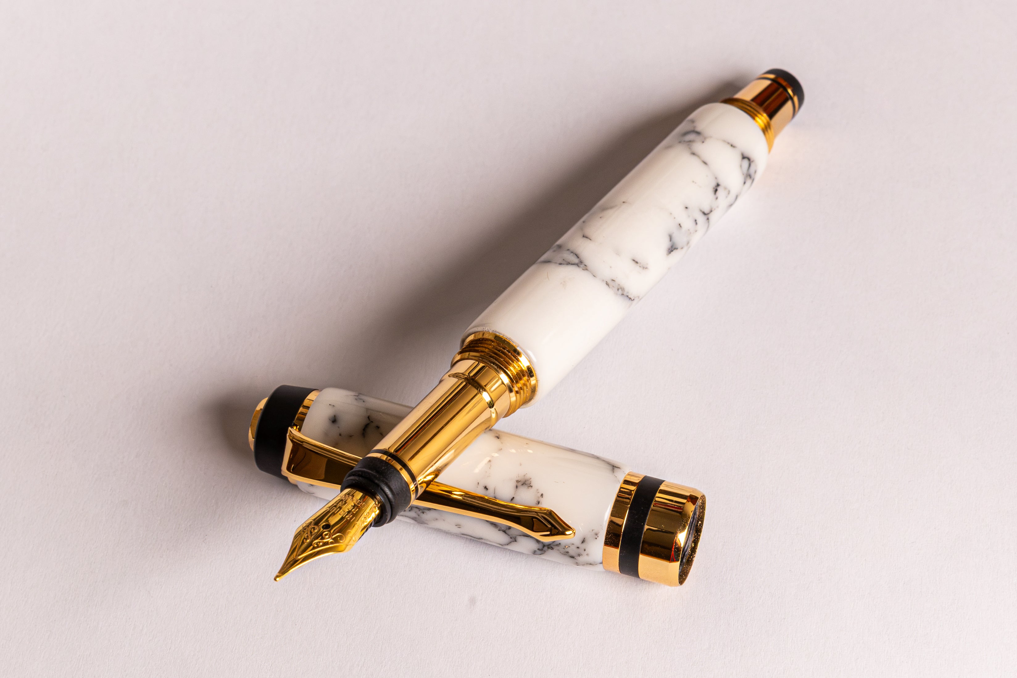 A luxury fountain pen with a white and gray marble-patterned body and polished gold-plated accents, shown with its cap removed and placed underneath the barrel on a light gray background.