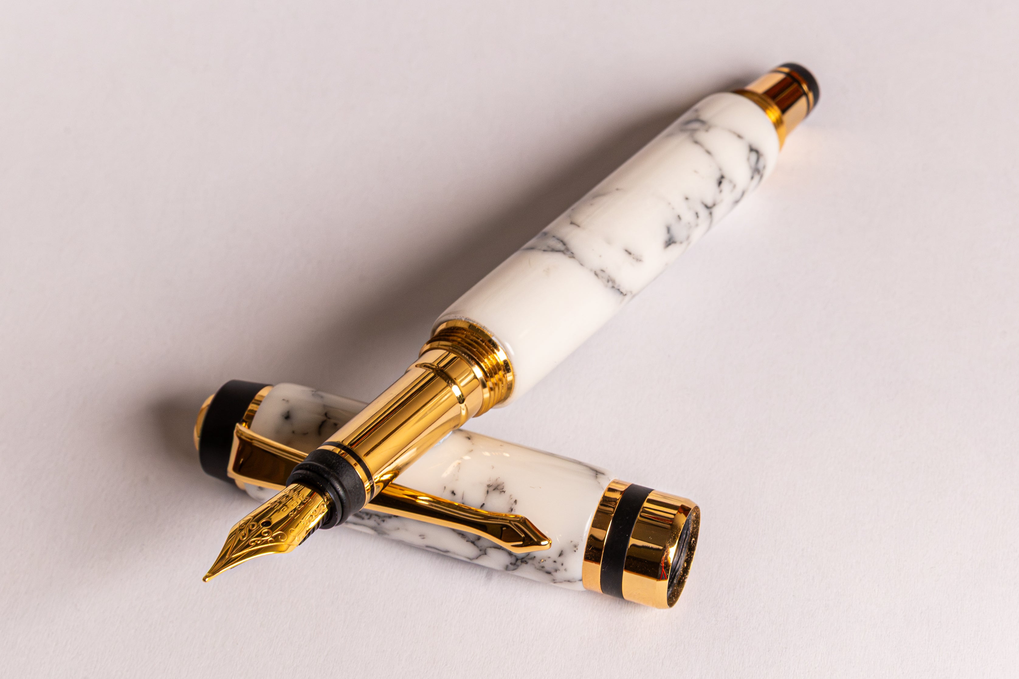 A luxury fountain pen with a white and gray marble barrel and cap, accented with shiny gold-plated metal trim and a gold nib, resting on a light gray background.