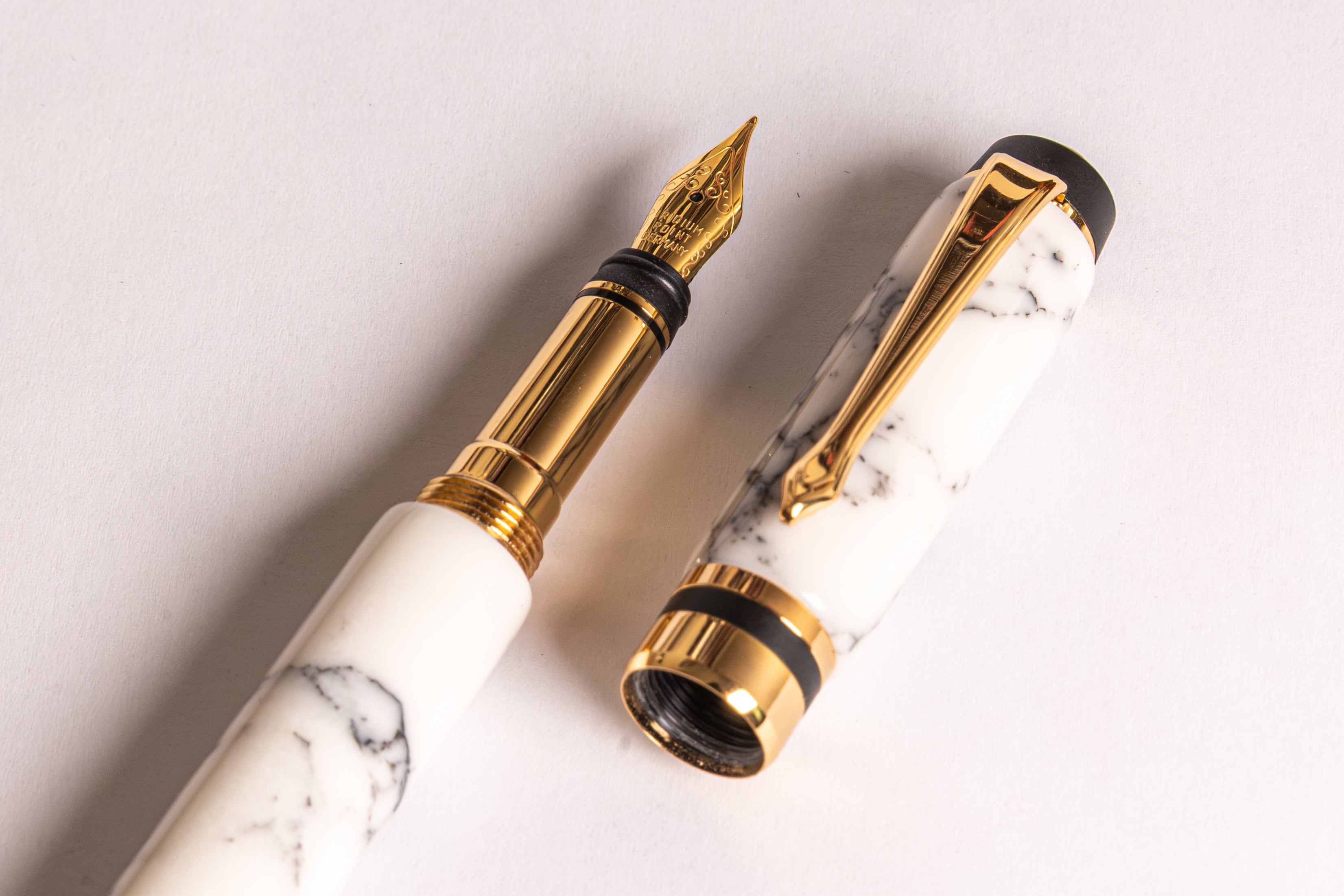 A close-up of a white marble fountain pen with gold trim and a gold nib, shown with its cap removed and placed beside the barrel on a light background.