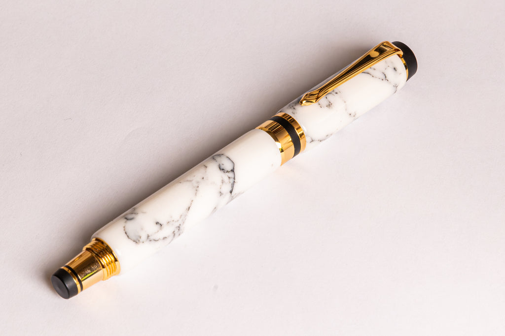 A luxury pen with a white and gray marble patterned barrel, accented by polished gold-tone metal trim and black details on the tip and base, resting diagonally on a clean white surface.
