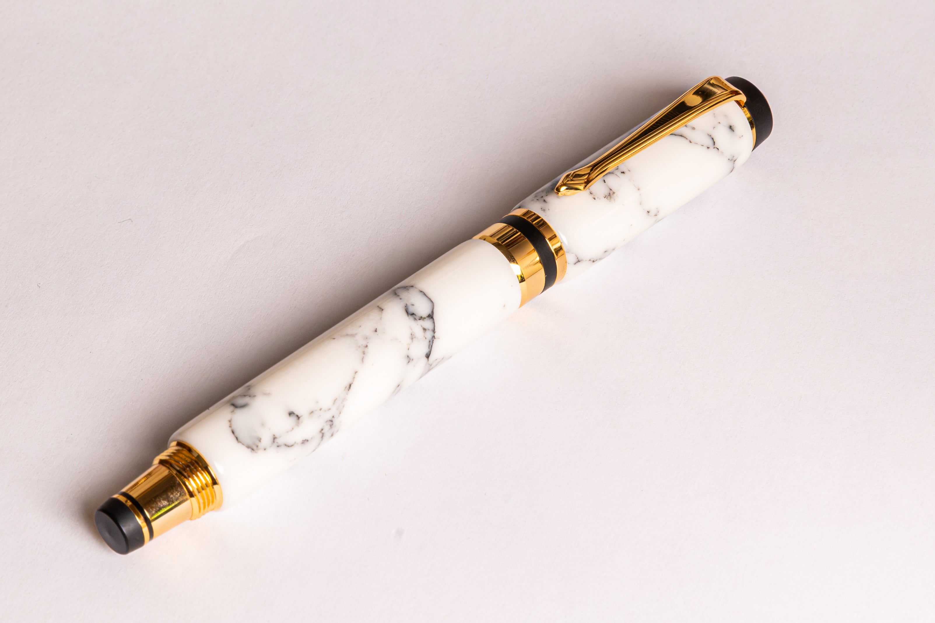 A luxury pen with a white and gray marble patterned barrel, accented by polished gold-tone metal trim and black details on the tip and base, resting diagonally on a clean white surface.