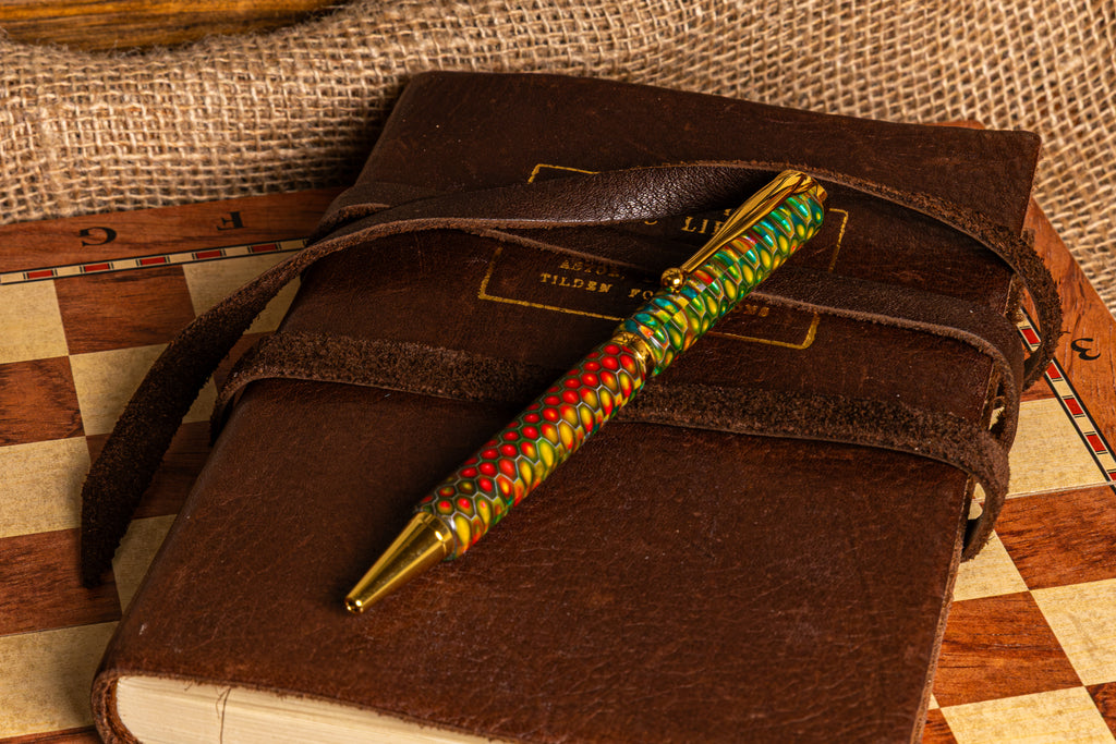 A decorative ballpoint pen with a colorful braided pattern in green, red, and yellow, accented with gold-toned metal details, resting on top of a dark brown leather journal.