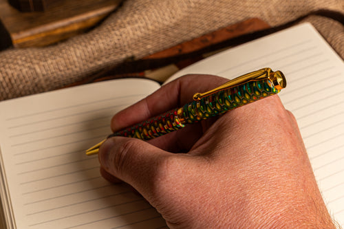 A close-up of a hand writing in an open lined notebook with a decorative pen. The pen has a gold-toned metal clip and tip, with a textured barrel featuring a multicolor pattern of green, gold, red, and blue.