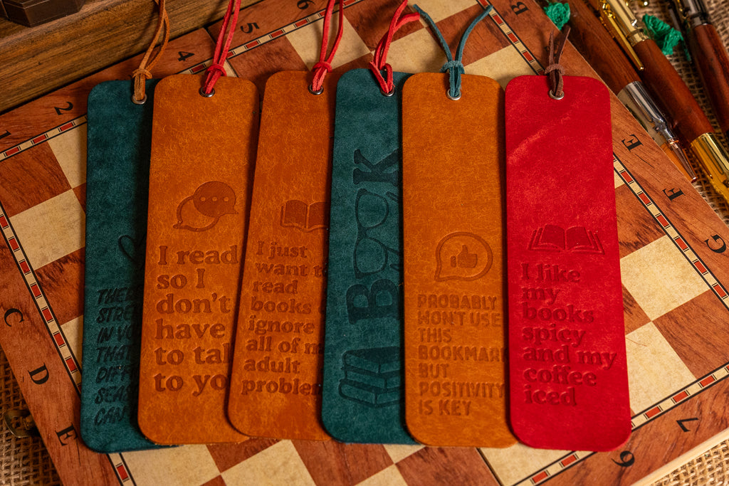 Six leather bookmark bookmarks in teal, tan, and red are lined up on a wooden chess board, each engraved with a positive book-themed quote and icon.
