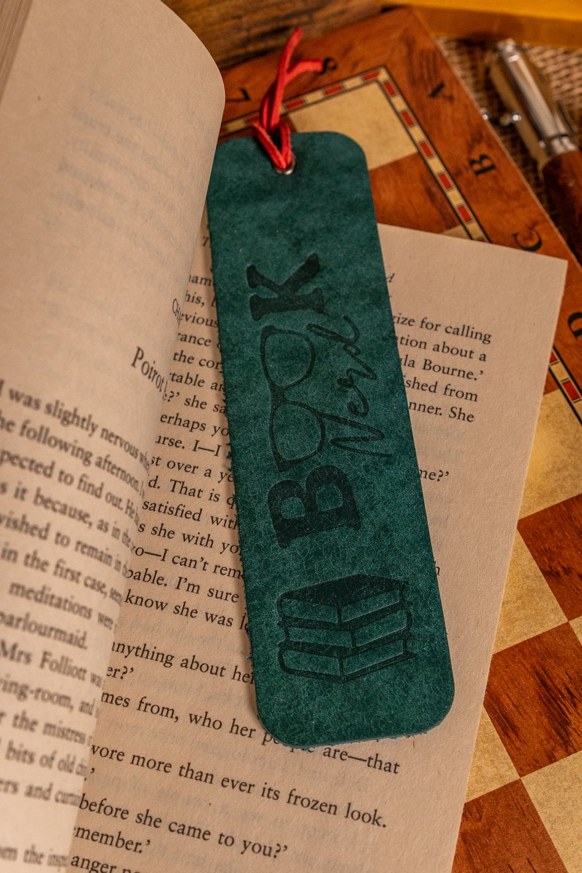 A dark teal green rectangular book tag with a red hanging string, featuring an embossed 'BOOK' and book spine design, resting on open book pages on a wooden table.